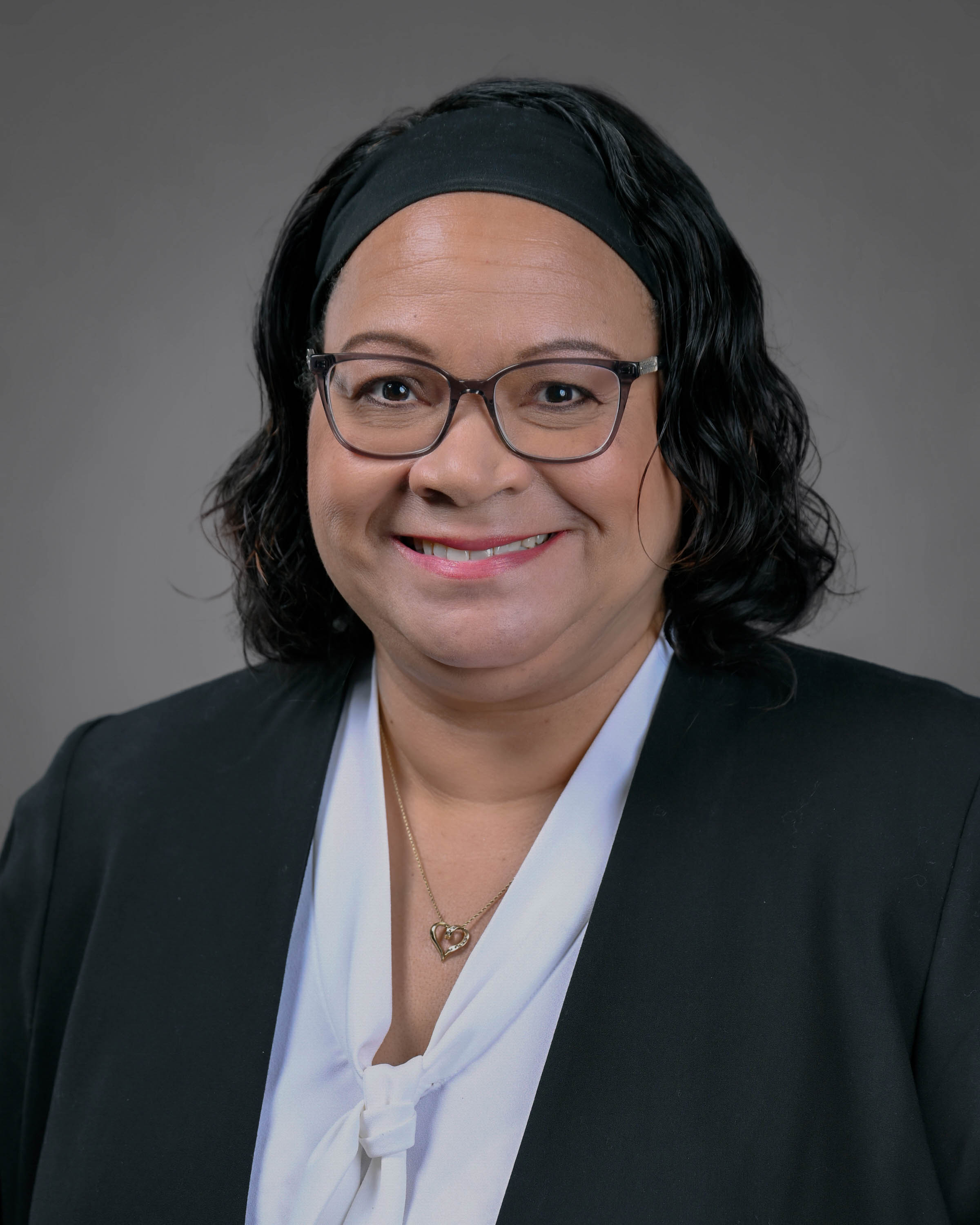 Professional headshot of a smiling woman with glasses and shoulder-length dark hair, wearing a black blazer and white blouse against a gray studio background.